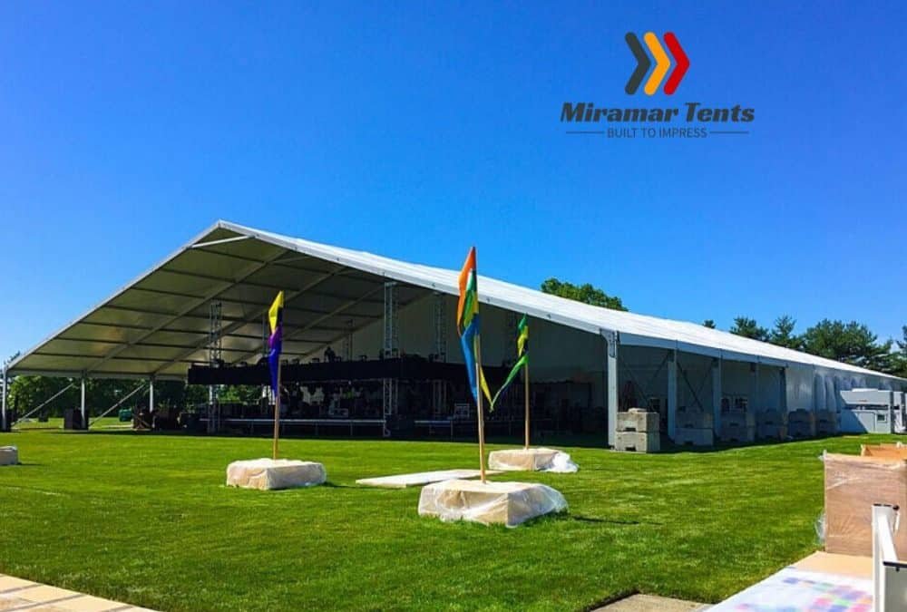 Large white All-Occasion Canopy Tent set up in a grassy area on a sunny day, with blue sky above and colorful flags at the entrance.