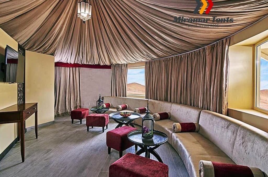 Luxurious desert tent interior furnished with plush sofas, an elegant glass table set with drinks, draped ceilings, and panoramic windows with expansive desert views.