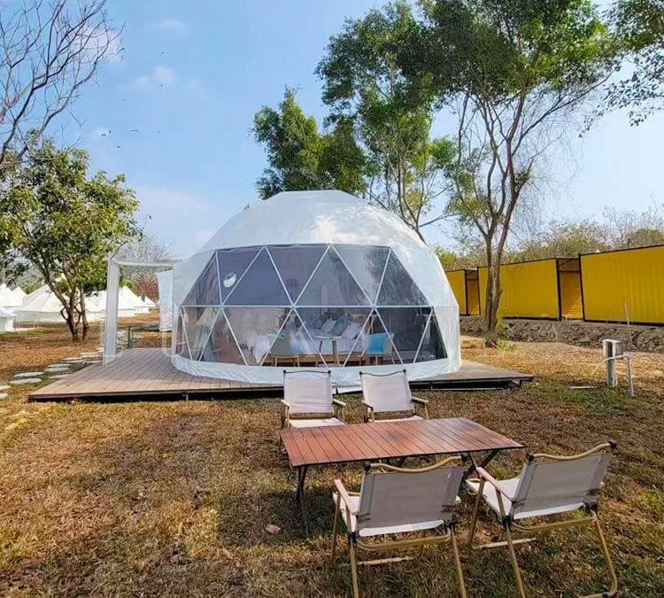 A geodesic dome tent on a wooden platform, surrounded by trees and grass, with a table and chairs in the foreground.