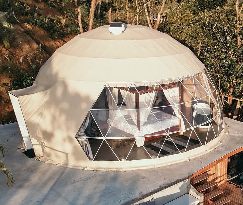 A dome-shaped glamping tent with a large geodesic window, revealing a bed inside. Surrounded by trees, the tent is set on an elevated platform with a small outdoor seating area.