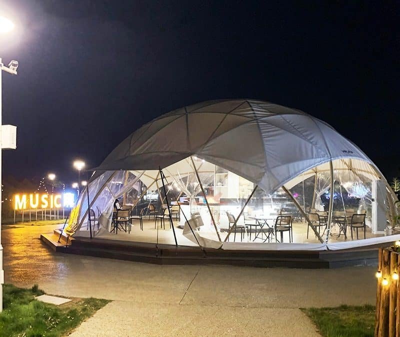 A geodesic dome structure with transparent panels is illuminated at night, housing tables and chairs inside. A large "MUSIC" sign is visible in the background to the left.