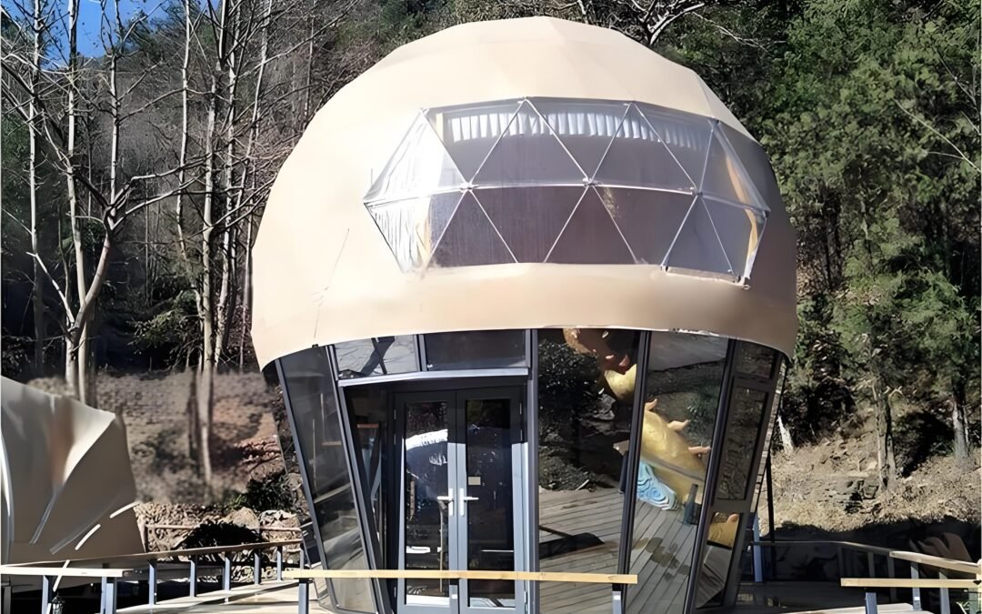 A dome-shaped structure with glass windows stands in a wooded area. It has a geodesic upper part and an entrance with double doors.