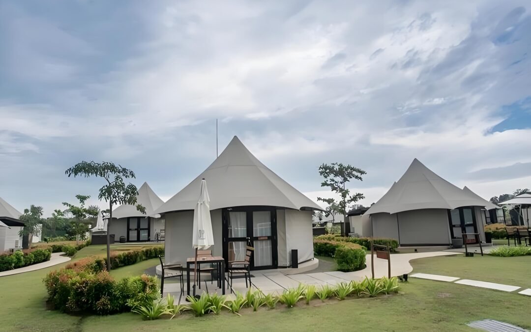 A peak tent hotel emerges as a cluster of conical tents on a grassy lawn, complete with a patio set. Surrounded by small shrubs and trees under a cloudy sky, this serene escape blends nature with comfort.