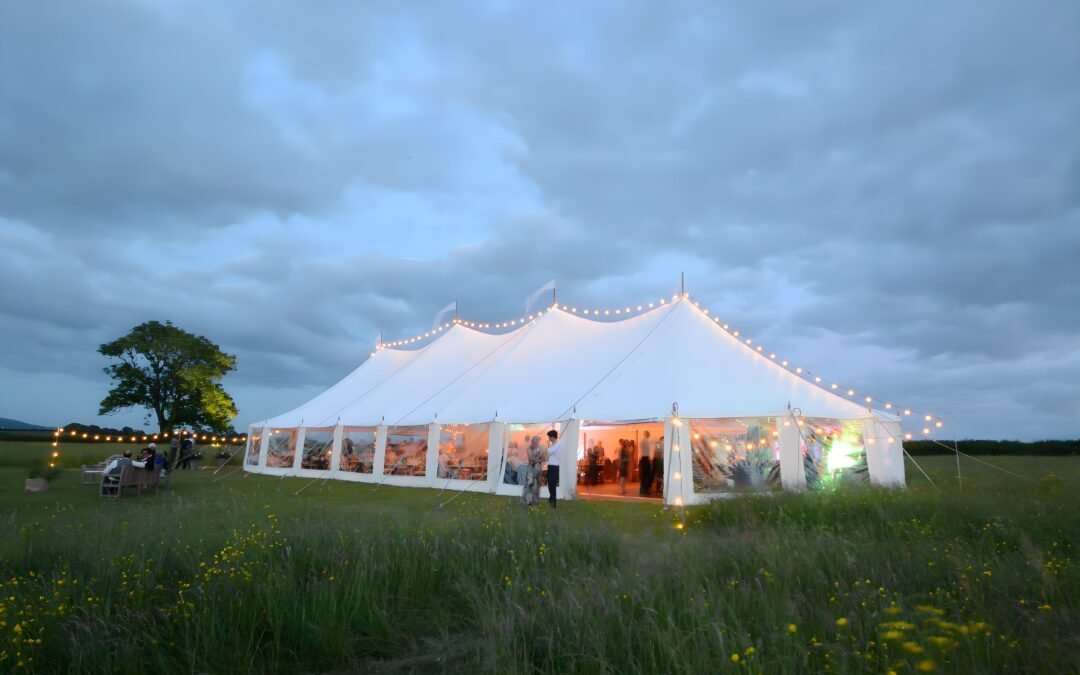 A large white party tent adorned with string lights stands in a grassy field under a cloudy sky, perfect for any event.