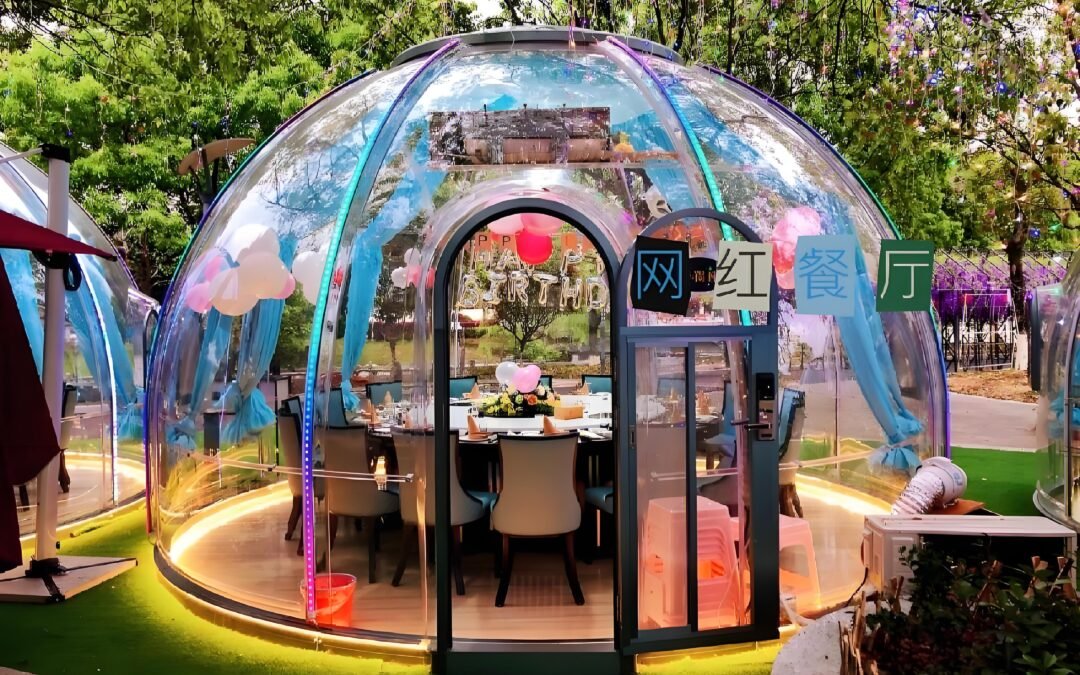 A transparent dining dome with clear walls, elegantly furnished with a table and chairs, is beautifully decorated with balloons and flowers and surrounded by lush greenery.