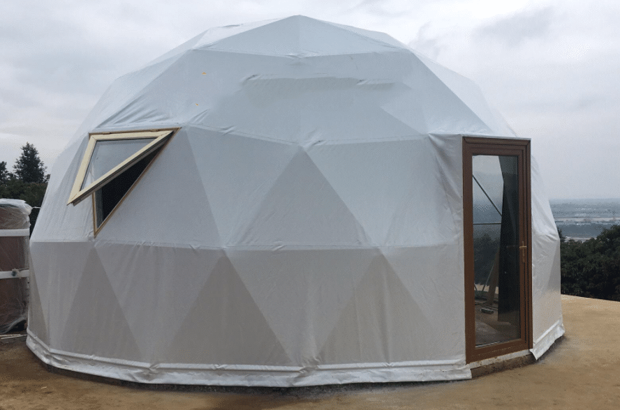 A glamping dome tent, covered with white material, features a wooden-framed glass door and a triangular window. It sits gracefully on a sandy surface under a cloudy sky.