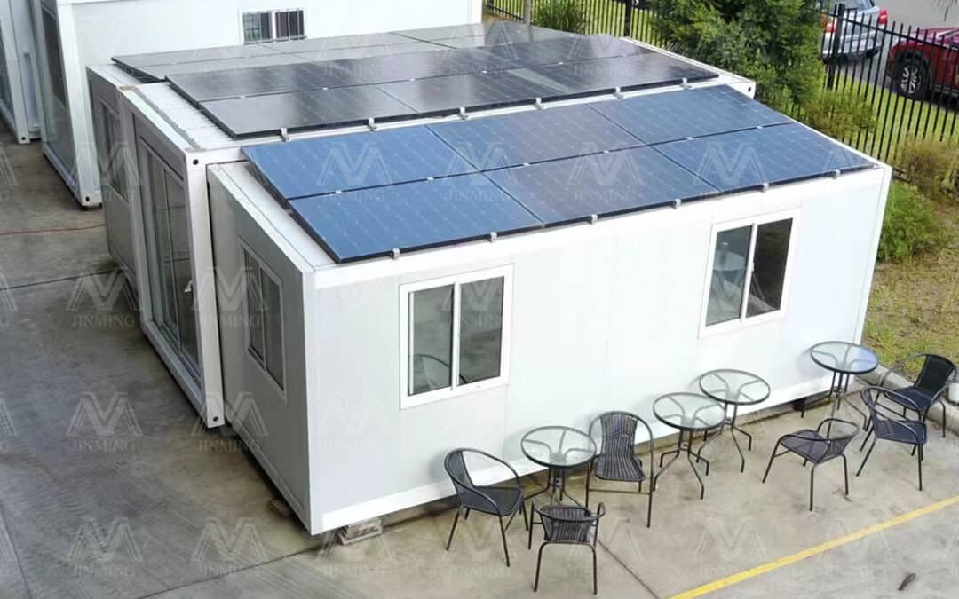 White Modular Building With Solar Panels On The Roof, Located On Concrete Ground. Five Outdoor Tables With Four Chairs Each Are Arranged Alongside The Building.
