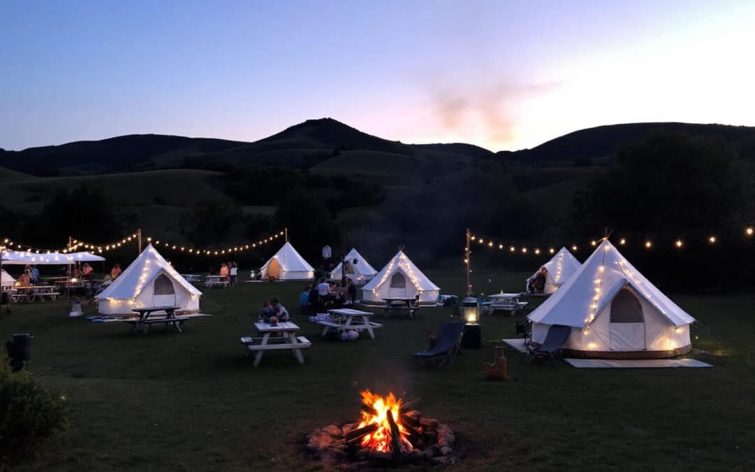 Glamping tents with string lights are set up in a field at dusk, with a campfire crackling in the center and hills silhouetted against the twilight sky.