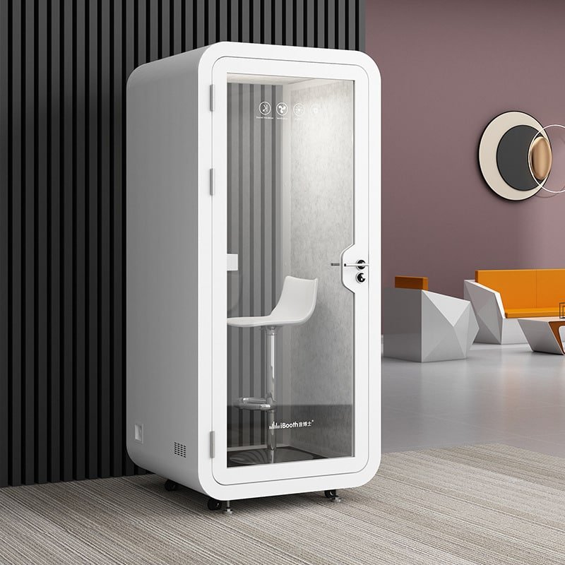 A white soundproof booth with a glass door and a white stool inside, positioned against a wall in a modern office space with geometric decor and furniture.