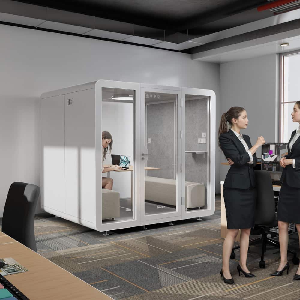 Two women in business attire talk near a glass walled office pod, where another person sits at a desk with a laptop in a modern open office environment.
