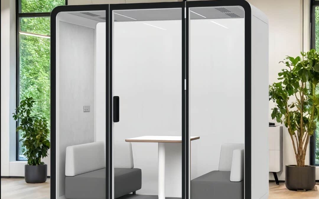 A modern office meeting pod with glass doors, two gray sofas, and a small white table inside, situated in a bright room with large windows and potted plants.